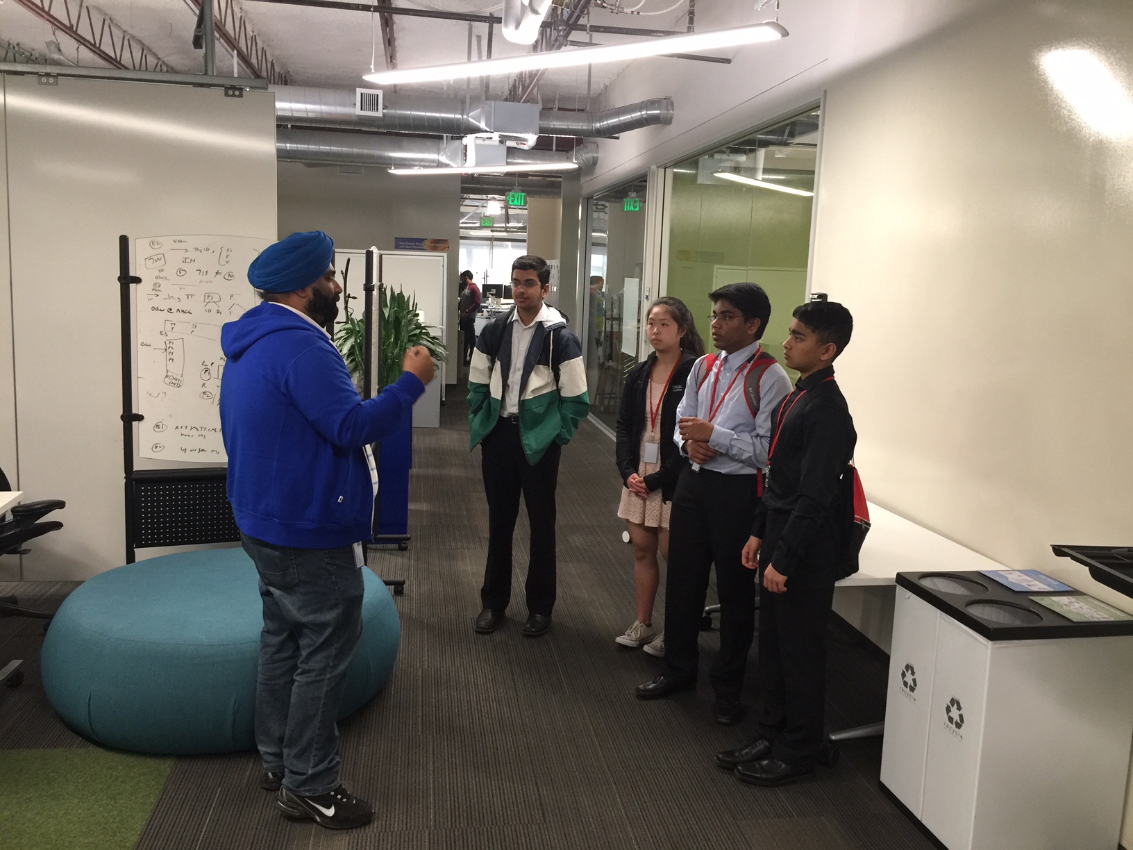 A group of students listening to a man speak in front of a whiteboard in an engineering company.